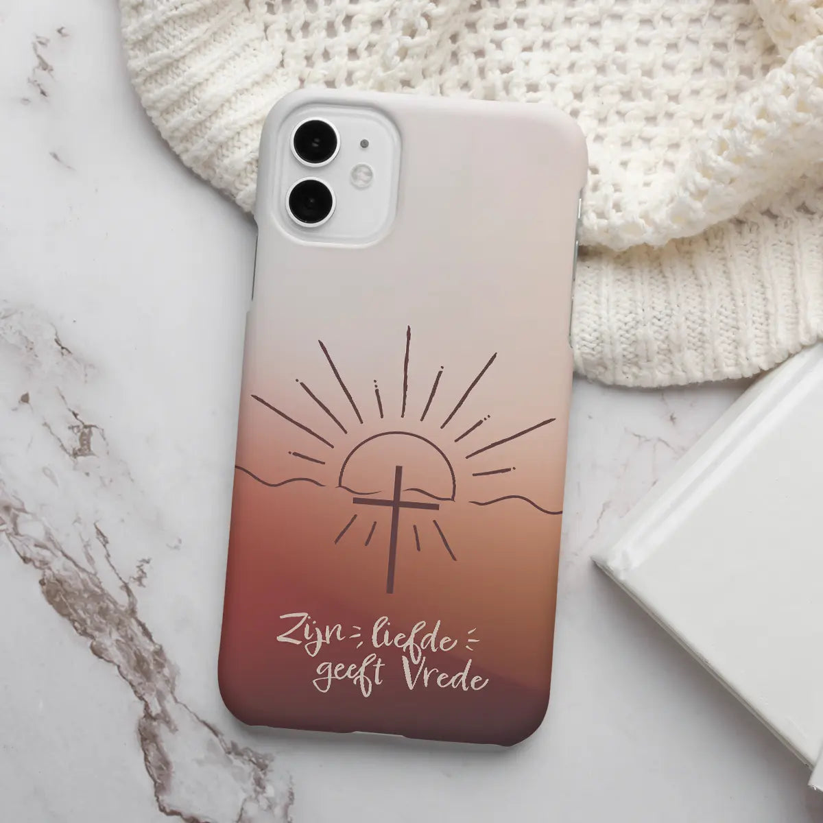 Hardcase christelijk telefoonhoesje met warm bruin verloop, fijn getekend kruis en opkomende zon, met de tekst ‘Zijn liefde geeft vrede’, liggend op een tafel in zacht natuurlijk licht.